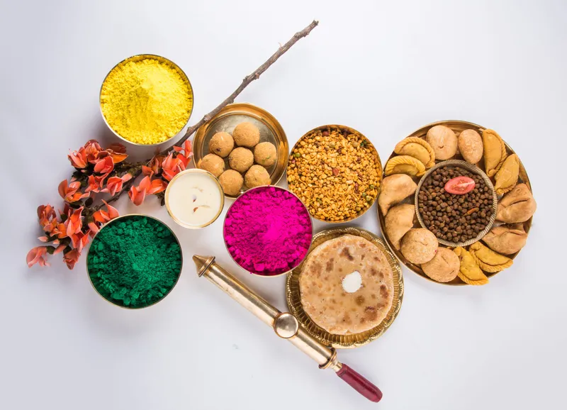 Happy Holi greeting card designed showing Indian traditional sweet and salty food, flowers and powder colours arranged over white background or clay. Selective focus