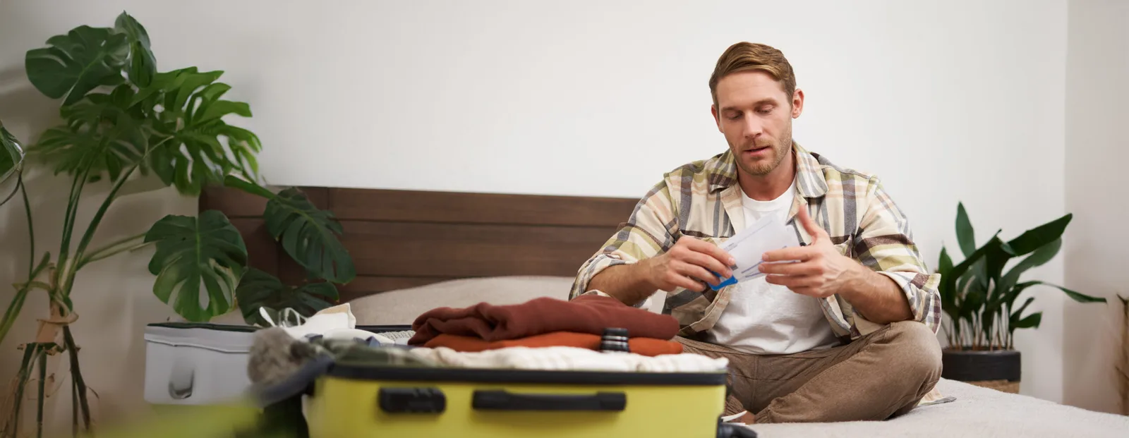 Portrait of handsome man, tourist, looking at his holiday tickets, booked vacation with travel agency, sitting on bed and packing clothes in suitcase